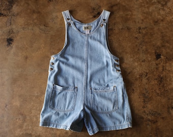 May include: A light blue denim overall romper featuring a scoop neckline, two front patch pockets, and metallic buttons along the sides and shoulder straps. The garment has a central vertical seam and a label at the neckline reading FRENZA.