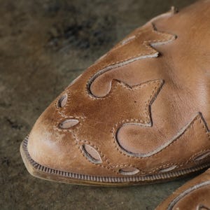 May include: Close-up of a tan leather boot with decorative cut-out and stitched star-like patterns. The boot has a rounded toe and a textured sole. The leather appears worn, giving it a vintage look.