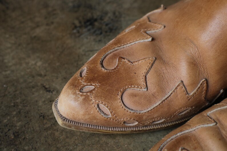 May include: Close-up of a tan leather boot with decorative cut-out and stitched star-like patterns. The boot has a rounded toe and a textured sole. The leather appears worn, giving it a vintage look.