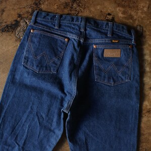 Puede incluir: Vaqueros de mezclilla azul oscuro con el logotipo de Wrangler en el bolsillo trasero. Los vaqueros tienen remaches y costuras de color cobre. Los bolsillos traseros presentan el dise&ntilde;o de costura "W" cl&aacute;sico de Wrangler.