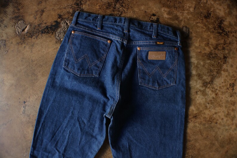 Puede incluir: Vaqueros de mezclilla azul oscuro con el logotipo de Wrangler en el bolsillo trasero. Los vaqueros tienen remaches y costuras de color cobre. Los bolsillos traseros presentan el dise&ntilde;o de costura "W" cl&aacute;sico de Wrangler.