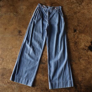 May include: Light blue denim pants with a high waist and wide legs. The pants have a button and zipper closure, with pleats at the front. The fabric has a slightly faded appearance, and the pants are laid flat on a brown surface.