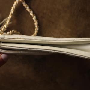 May include: Ivory-colored quilted shoulder bag with a gold chain strap and a tassel. The bag is rectangular with a flap closure. The chain is intertwined with a white material. The bag is held by a hand.