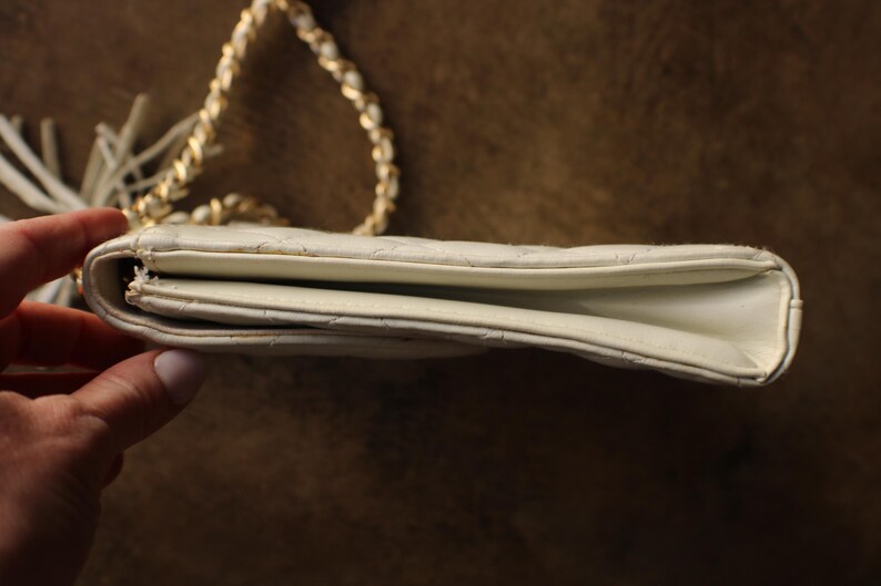 May include: Ivory-colored quilted shoulder bag with a gold chain strap and a tassel. The bag is rectangular with a flap closure. The chain is intertwined with a white material. The bag is held by a hand.