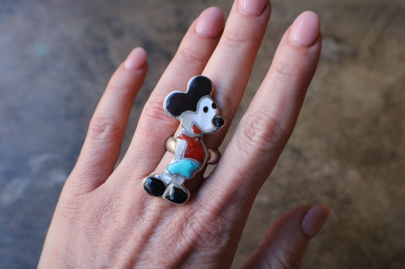 Peut inclure: Une bague en argent avec un motif incrust&eacute; de plusieurs pierres en forme de souris de dessin anim&eacute;. L'incrustation se compose de pierres noires, blanches, rouges et turquoise serties dans une monture en argent poli, formant une figurine.