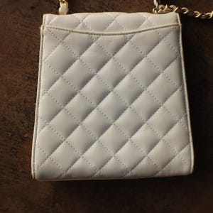 May include: A small, white quilted handbag with a gold chain strap. The bag has a rectangular shape with a flap closure and a diamond pattern. The background is a dark brown wooden surface.