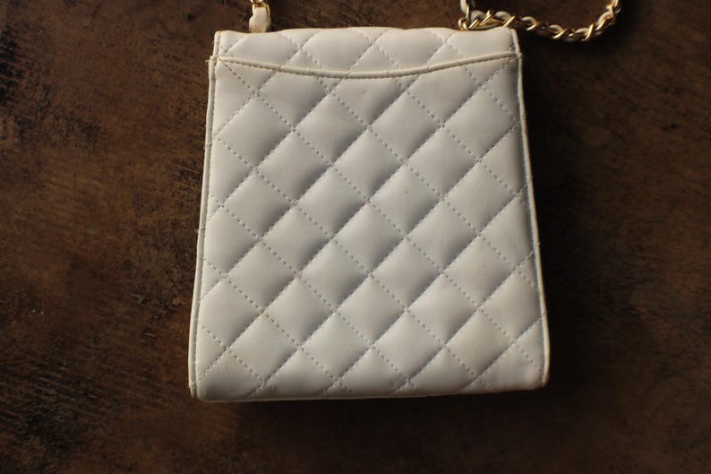 May include: A small, white quilted handbag with a gold chain strap. The bag has a rectangular shape with a flap closure and a diamond pattern. The background is a dark brown wooden surface.