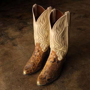 May include: A pair of cowboy boots with a cream-colored upper featuring decorative stitching and a brown snakeskin lower. The boots have a pointed toe and a gold-colored metal accent on the toe.