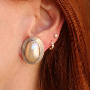 Puede incluir: Primer plano de una oreja con tres pendientes. El pendiente más grande es un pendiente de plata ovalado con una perla grande de color crema. También se ven un pequeño diamante y un pequeño pendiente de aro.