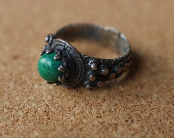 Puede incluir: Un anillo de plata con una piedra preciosa redonda de malaquita verde pulida en un engaste tipo corona. La banda está hecha de plata oscura oxidada con una superficie texturizada y abultada, con detalles metálicos intrincados alrededor de la piedra.