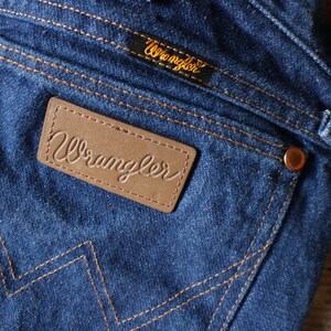 Puede incluir: Primer plano de vaqueros de mezclilla azul con remaches y costuras cobrizas. El bolsillo trasero presenta un parche de cuero marr&oacute;n con la palabra "Wrangler" en relieve. Tambi&eacute;n es visible una peque&ntilde;a etiqueta negra con "Wrangler" en letras doradas.