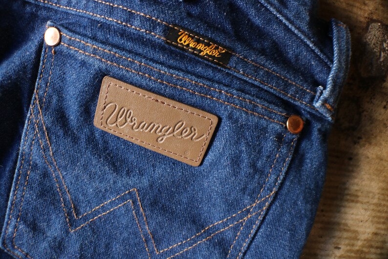 Puede incluir: Primer plano de vaqueros de mezclilla azul con remaches y costuras cobrizas. El bolsillo trasero presenta un parche de cuero marr&oacute;n con la palabra "Wrangler" en relieve. Tambi&eacute;n es visible una peque&ntilde;a etiqueta negra con "Wrangler" en letras doradas.