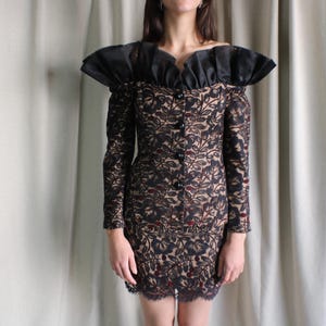 May include: A vintage black lace dress with a floral design, long sleeves, and a ruffled neckline. The dress has a button-down front and a matching skirt. The hemline is trimmed with lace.