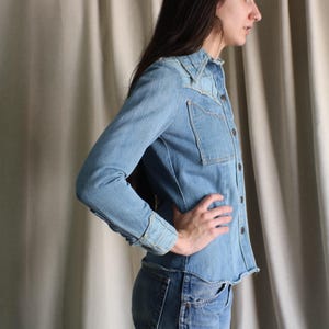 May include: Light blue denim shirt with long sleeves, a collared neckline, and button-down front. The shirt features two chest pockets and is paired with matching denim jeans. The outfit is a classic, casual style.