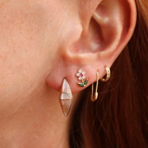 Puede incluir: Primer plano de una oreja adornada con múltiples pendientes. Un pendiente dorado, alargado en forma de lágrima, cuelga del lóbulo. Encima, un pequeño pendiente de flor con pétalos blancos y centro rojo. Un imperdible dorado y un pequeño pendiente de aro completan el conjunto.