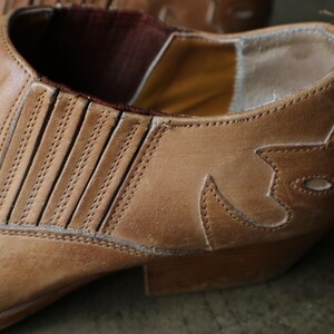 May include: Close-up of a tan leather boot with decorative stitching and a pointed toe. The boot features a western-style design with a low heel and elastic side panels for easy wear. The leather has a worn appearance.