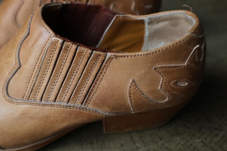 May include: Close-up of a tan leather boot with decorative stitching and a pointed toe. The boot features a western-style design with a low heel and elastic side panels for easy wear. The leather has a worn appearance.