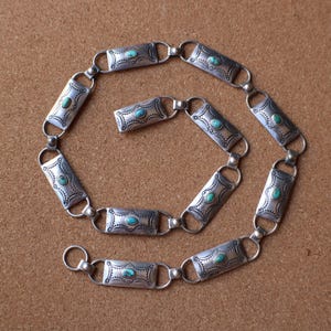 以下が含まれることがあります： 長方形のリンクが付いたシルバーカラーのベルト。各リンクにはターコイズ色の石が使用されています。リンクは小さなリングで接続されています。ベルトは茶色の表面に表示されています。