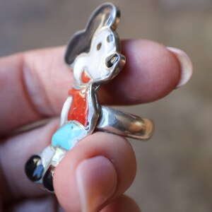 Peut inclure: Une bague en argent avec un motif de personnage de dessin anim&eacute; incrust&eacute;. La figurine pr&eacute;sente un visage blanc, des oreilles noires, un haut rouge, un short bleu clair et des chaussures noires, le tout serti dans une monture en argent poli.
