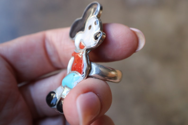 Peut inclure: Une bague en argent avec un motif de personnage de dessin anim&eacute; incrust&eacute;. La figurine pr&eacute;sente un visage blanc, des oreilles noires, un haut rouge, un short bleu clair et des chaussures noires, le tout serti dans une monture en argent poli.