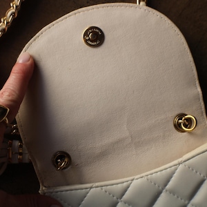 May include: Cream-colored quilted handbag with a gold chain strap and gold hardware. The interior is a lighter cream color. The bag has a flap closure with a gold snap. The bag is open, revealing the interior.