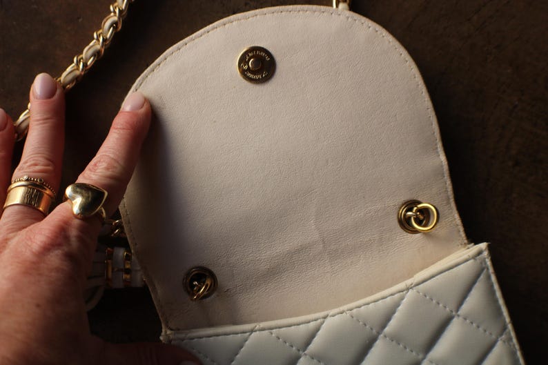 May include: Cream-colored quilted handbag with a gold chain strap and gold hardware. The interior is a lighter cream color. The bag has a flap closure with a gold snap. The bag is open, revealing the interior.
