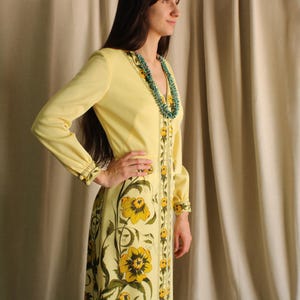 Könnte beinhalten: Ein gelbes Vintage-Kleid mit langen Ärmeln und V-Ausschnitt. Das Kleid hat ein Blumenmuster in Gelb und Grün, mit einem passenden Design auf der Vorderseite und den Ärmeln. Eine türkisfarbene Halskette setzt einen farblichen Akzent.