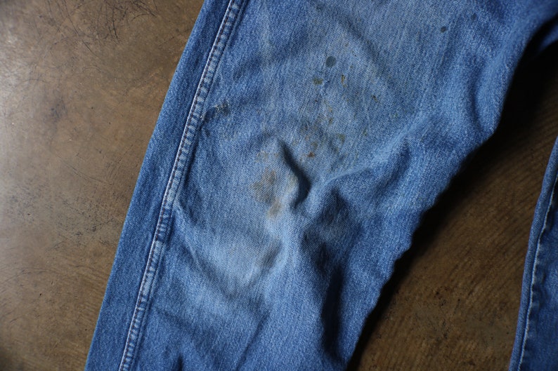 Puede incluir: Primer plano de unos vaqueros de mezclilla azul descoloridos. Los vaqueros muestran signos de desgaste, con manchas y decoloraci&oacute;n visibles en la tela. Las costuras son claramente visibles.