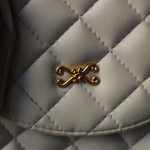 May include: Close-up of a white quilted handbag with a gold-colored decorative clasp. The bag features a diamond pattern and a tassel detail on the left side. The clasp is an ornate, symmetrical design.