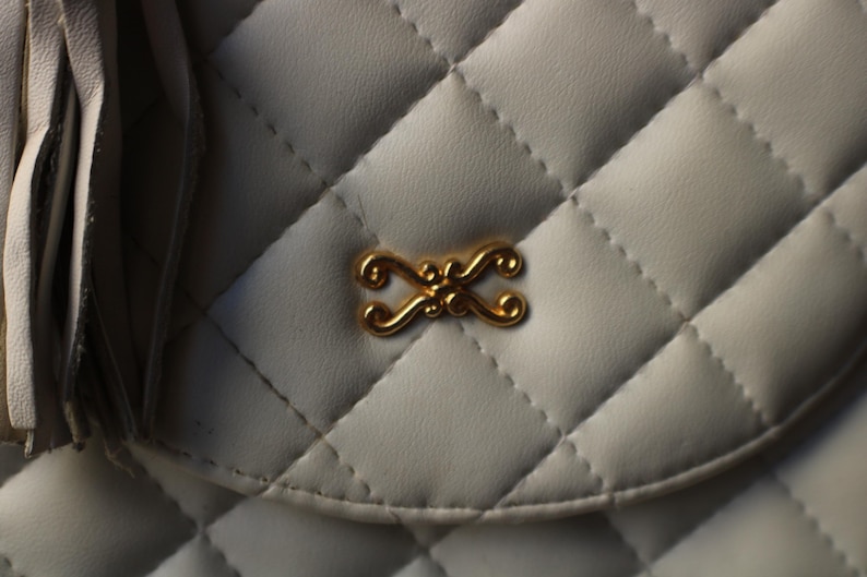 May include: Close-up of a white quilted handbag with a gold-colored decorative clasp. The bag features a diamond pattern and a tassel detail on the left side. The clasp is an ornate, symmetrical design.