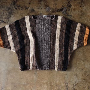 May include: A striped, short-sleeved sweater in shades of brown, black, and cream. The sweater has a textured appearance and a relaxed fit. The sleeves have a slight orange accent at the ends. The garment is displayed on a neutral surface.
