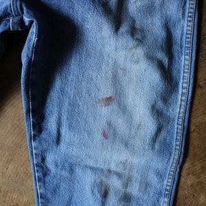 Puede incluir: Primer plano de unos vaqueros de mezclilla azul claro con desgaste visible. Los vaqueros tienen un dise&ntilde;o cl&aacute;sico de cinco bolsillos y muestran signos de manchas y decoloraci&oacute;n, lo que se suma a su est&eacute;tica vintage. La imagen resalta la textura y los detalles de la tela vaquera.
