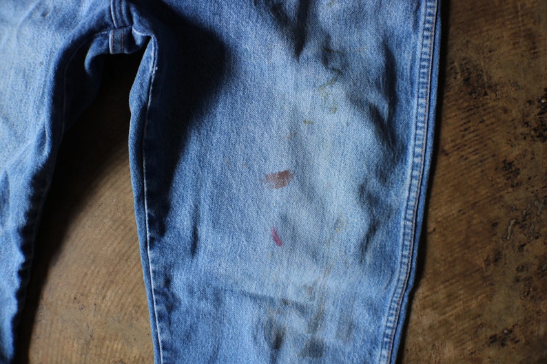 Puede incluir: Primer plano de unos vaqueros de mezclilla azul claro con desgaste visible. Los vaqueros tienen un dise&ntilde;o cl&aacute;sico de cinco bolsillos y muestran signos de manchas y decoloraci&oacute;n, lo que se suma a su est&eacute;tica vintage. La imagen resalta la textura y los detalles de la tela vaquera.