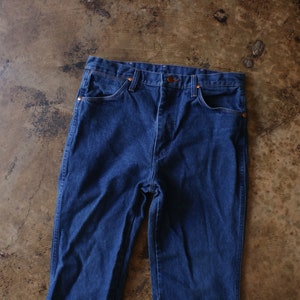 May include: A pair of dark blue denim jeans featuring a high-waisted design, a button fly closure, and copper-toned rivets at the corners of the front pockets. The jeans have a classic five-pocket construction with visible stitching along the seams.