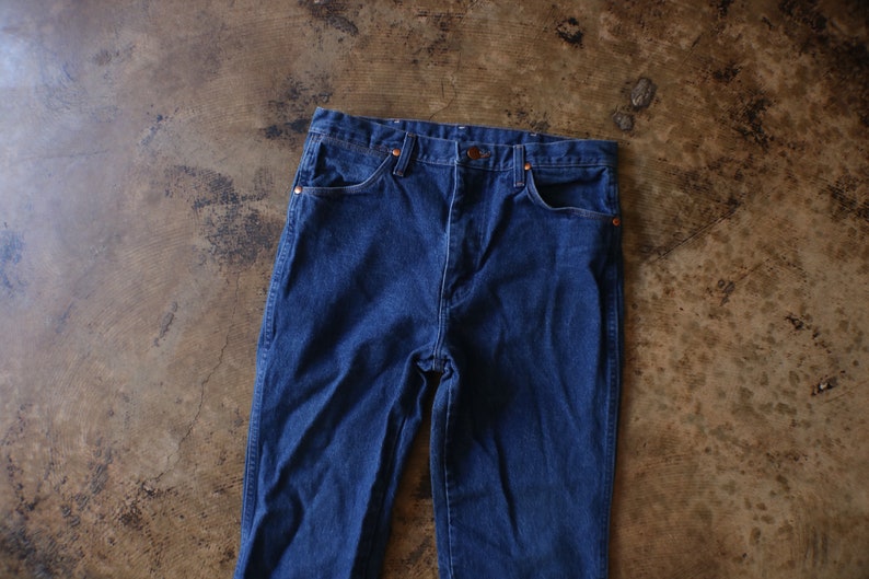 May include: A pair of dark blue denim jeans featuring a high-waisted design, a button fly closure, and copper-toned rivets at the corners of the front pockets. The jeans have a classic five-pocket construction with visible stitching along the seams.
