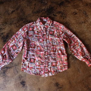 May include: A long-sleeved button-down shirt featuring a geometric design in red, brown, and white. The shirt has a collared neckline and is displayed flat. A vintage-style garment.