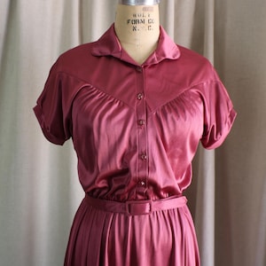 May include: A short-sleeved, button-down dress in a solid dark rose color. The garment features a pointed collar, a V-shaped yoke across the chest, and a matching fabric belt at the waist. The material has a smooth, satin-like sheen throughout.