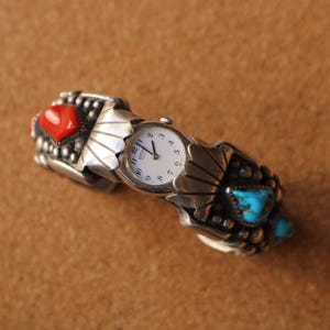 May include: Silver-toned watch with a white face and black numerals. The watch band features red and turquoise stones, and a decorative design. The watch is set against a brown background.