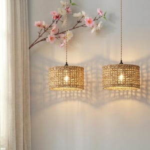 May include: Two woven pendant lights with cylindrical shades suspended from the ceiling. The light brown shades have a grid-like pattern. The lights are on, creating shadows on the wall. A branch with pink and white flowers is in the background.
