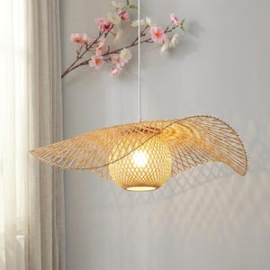 May include: A woven bamboo pendant light fixture. The light has a spherical, woven shade with a bulb inside, and a wide, hat-like shade around it. The light hangs from a white cord. The background includes a curtain and a branch with pink flowers.
