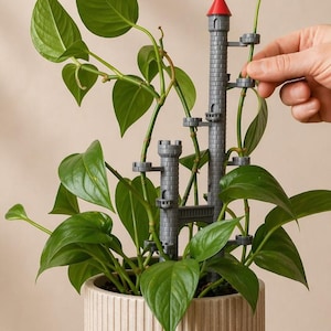 May include: A potted plant with vibrant green leaves, featuring a gray castle-shaped plant support with a red-tipped tower. The plant is in a beige, ribbed ceramic pot. A hand is adjusting the plant's vines around the castle structure.