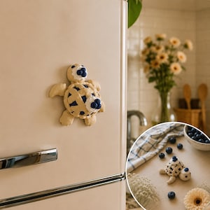 May include: A whimsical, beige turtle-shaped magnet with a pie crust design on its shell, adorned with blue berry accents. The magnet is attached to a cream-colored refrigerator. A smaller version of the turtle is on a table with blueberries.