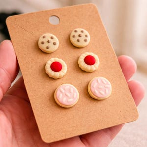 May include: A selection of six cookie-shaped stud earrings presented on a brown card. The earrings showcase designs such as chocolate chip, jam-filled, and pink iced cookies. The earrings are small and colourful.