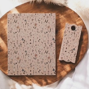 May include: A notebook and phone case with a beige background and a repeating pattern of geese, flowers, and foliage. The notebook is rectangular, and the phone case has a hole for the camera. Both items are on a wooden tray.
