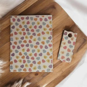 May include: A notebook and a phone case featuring a pattern of colourful candy hearts on a cream background. The hearts are in pastel shades and have short phrases. The items are on a wooden surface.