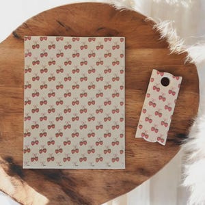 May include: A notebook and bookmark with a repeating pattern of red cherries on a cream background. The notebook is rectangular, and the bookmark has a curved bottom with a hole at the top. The items are on a wooden tray.