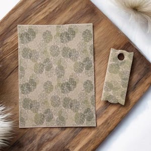 May include: A rectangular paper with a floral pattern and a small, matching tag with a hole. The design features a repeating pattern of green and gray flowers on a beige background. The items are on a wooden surface.