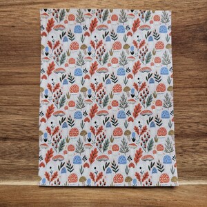 May include: A rectangular fabric with a repeating pattern of mushrooms, leaves, and berries in shades of red, orange, blue, and green on a white background. The fabric is displayed on a wooden surface.