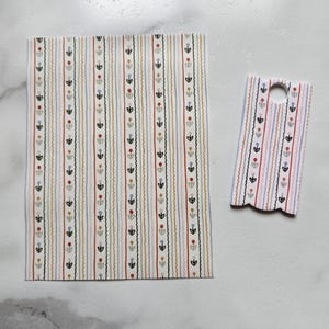 May include: Two pieces of patterned paper with a floral and striped design. The larger piece is rectangular, and the smaller piece has a hole at the top. The pattern includes vertical stripes in various colors, including red, blue, and yellow.
