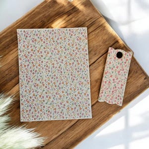 May include: A rectangular notebook and a small cutting board with a floral pattern of pink, purple, and yellow flowers on a cream background. The items are on a wooden surface.